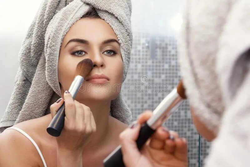 Помогает макияжу Woman with a Towel on Her Head, Looking in the Mirror and Applying Make Up Stock